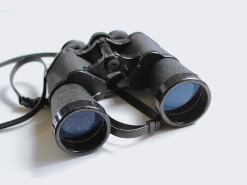 A pair of black binoculars with textured grips and blue lenses, resting on a light background.
